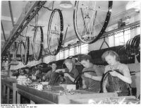 "Zwei Frauenbrigaden fädeln in zwei Schichten hier mit flinken Händen die Speichen ein. Die fertigen Räder werden an einen Ringtransporteur gehängt, der an der Decke entlangläuft und die Räder zur weiteren Bearbeitung in die anderen Räume transportiert." (April 1953)