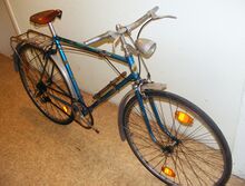 Sportrad im Fundzustand. Die Naben sind von 1955 und 1956. Letzteres als Baujahr angenommen, sind Scheinwerfer, Bremsen, Sattel, Luftpumpe und Bereifung offensichtlich nachgerüstet. Verbaut ist eine Rennrad-Kurbelgarnitur. Laufradgröße 28 Zoll.