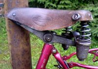 Tourensattel von ?? Zeitraum: 195? bis 1957 Verwendung: Simson-Fahrräder Decke: Leder, lackiert Gestell: Stahl, verchromt Bemerkungen: Ausführungen für Damen- und Herrenräder, Regulierfedern, mit Simson-Plakette aus Blech an der Rückseite, Hersteller unbekannt