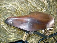 Tourensattel von ?? Zeitraum: 195? bis 1957 Verwendung: Simson-Fahrräder Decke: Leder, lackiert Gestell: Stahl, verchromt und lackiert Bemerkungen: Ausführungen für Damen- und Herrenräder, genäht, mit Simson-Plakette an der Rückseite, Hersteller unbekannt